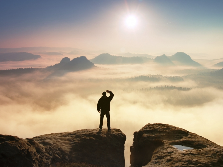 Colorful misty morning in rocks. Tourist in dark cloths on rock empire shadowing eyes  with hand in the air.